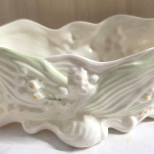May include: A cream-colored ceramic bowl featuring a sculpted relief of lily of the valley flowers and leaves with soft green accents. The bowl has an irregular, wavy rim and a base formed by rounded, organic shapes with a glossy finish.