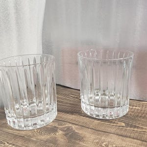 以下が含まれることがあります： 2つの透明なリブガラスのタンブラー。グラスは、垂直線と頑丈なベースを備えたクラシックなデザインです。飲み物の提供に適しています。グラスは木製の表面に表示されています。
