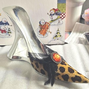 May include: A decorative silver high-heeled shoe with a leopard print toe and a black lace and feather accent. The shoe features an orange gem and is set against a festive winter-themed background.