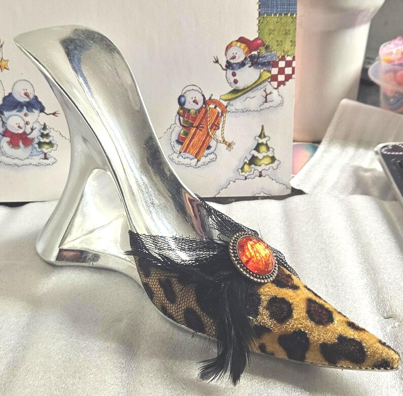 May include: A decorative silver high-heeled shoe with a leopard print toe and a black lace and feather accent. The shoe features an orange gem and is set against a festive winter-themed background.