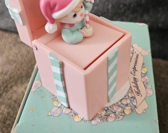 Op de afbeelding: Een roze cadeaudoos met een wit en groen gestreept ontwerp. De doos is open en er zit een klein beeldje van een kind met een kerstmanmuts in. De doos staat op een groene en witte doos met de woorden "Holiday Expressions" en "The Enesco Precious Moments Collection" erop.