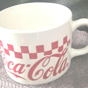 Puede incluir: Taza de Coca-Cola de cerámica blanca con un patrón de cuadros rojos y el logotipo de Coca-Cola. La taza tiene un asa curva y una abertura ancha. Para bebidas.
