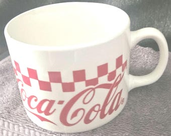 Op de afbeelding: Witte keramische Coca-Cola-mok met een rood geblokt patroon en het Coca-Cola-logo. De mok heeft een gebogen handvat en een brede opening. Voor dranken.
