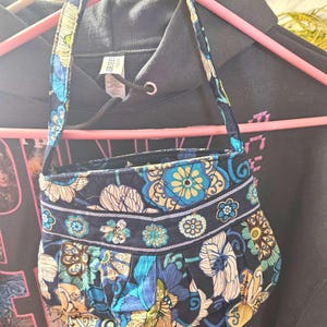 May include: A small, quilted handbag with a floral pattern in shades of blue, yellow, and white. The bag has a matching strap and is hanging on a pink hanger. The background is a dark gray hoodie.