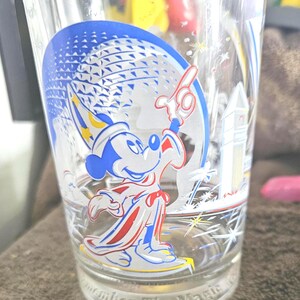 May include: Clear glass featuring a blue and white graphic of Mickey Mouse in a sorcerer's outfit, with a blue and white sphere in the background. The glass has the text "Remember The Magic" at the base. A decorative item.