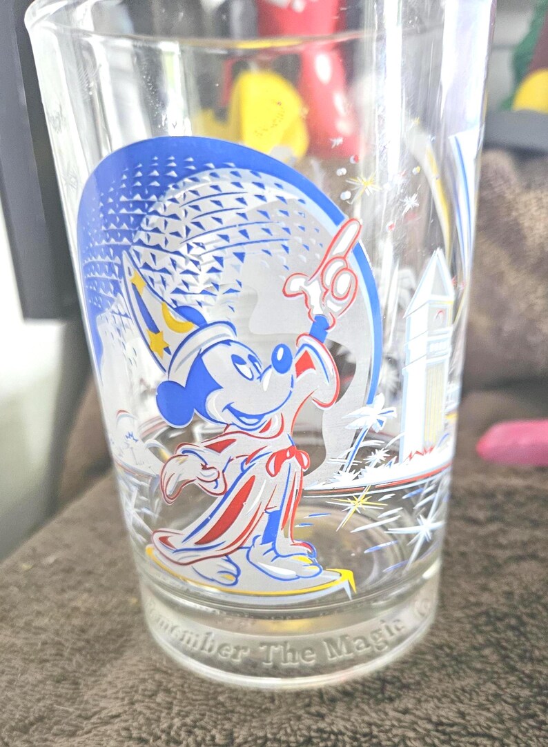 May include: Clear glass featuring a blue and white graphic of Mickey Mouse in a sorcerer's outfit, with a blue and white sphere in the background. The glass has the text "Remember The Magic" at the base. A decorative item.