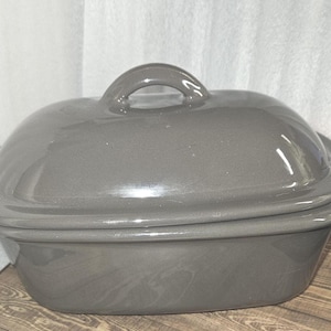 May include: A gray oval-shaped ceramic casserole dish with a matching lid and a rounded handle. The dish has a glossy finish and sits on a wooden surface. The background is a white curtain.