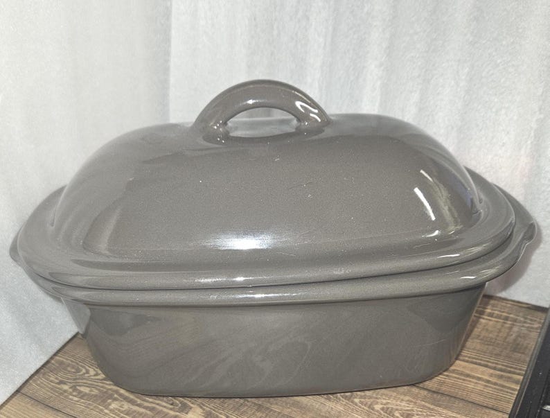 May include: A gray oval-shaped ceramic casserole dish with a matching lid and a rounded handle. The dish has a glossy finish and sits on a wooden surface. The background is a white curtain.