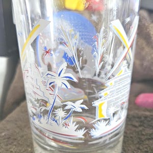 May include: Clear glass with a decorative design featuring palm trees, fireworks, and architectural elements. The glass has a rounded base and a straight, slightly flared top. The words "Walt Disney" are visible at the base.