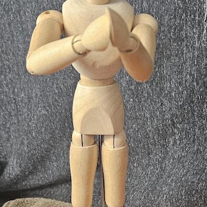 May include: A wooden articulated figure with jointed arms, legs, and head. The figure is standing with its arms crossed in front of its chest. The figure is on a wooden base with the word "Heson" printed on it.