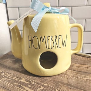 May include: A pale yellow ceramic teapot-shaped birdhouse with a handle, a spout, and a circular opening in the front. The word HOMEBREW is printed in black lettering on the front. A light blue ribbon is tied around the top, with a tag reading BY MAGENTA.