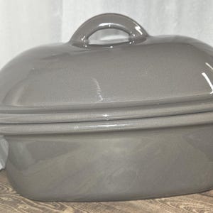 May include: A gray oval-shaped ceramic casserole dish with a lid and a handle. The dish is sitting on a wooden surface. The lid has a rounded shape and a small loop handle.