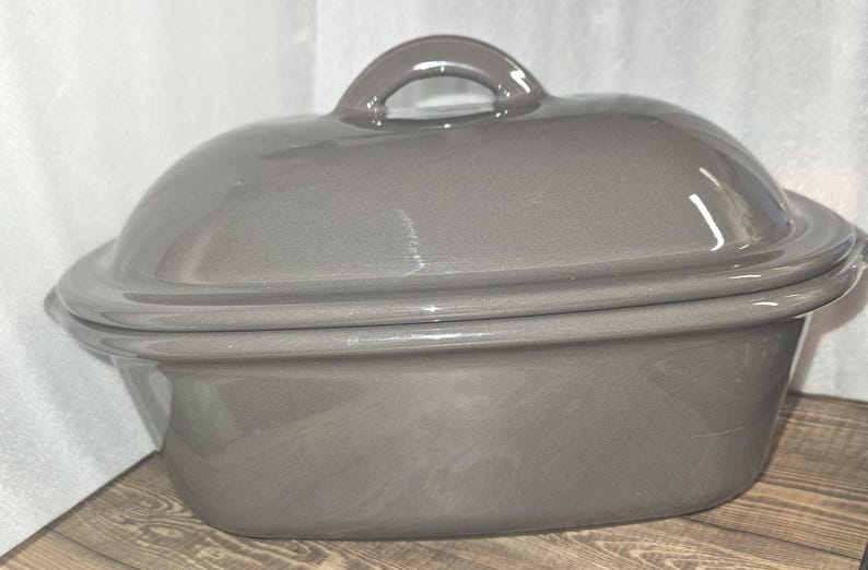 May include: A gray oval-shaped ceramic casserole dish with a lid and a handle. The dish is sitting on a wooden surface. The lid has a rounded shape and a small loop handle.
