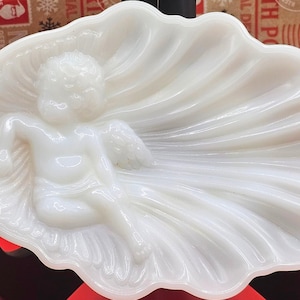May include: A white, opalescent glass dish in the shape of a seashell. A cherubic figure with wings reclines within the shell. The dish has a glossy finish and is likely vintage.