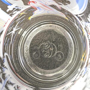 May include: Clear glass with a decorative rim featuring white starbursts and a colorful design. The bottom of the glass has the text "Remember the Magic" and a carriage with the number "25" inside.
