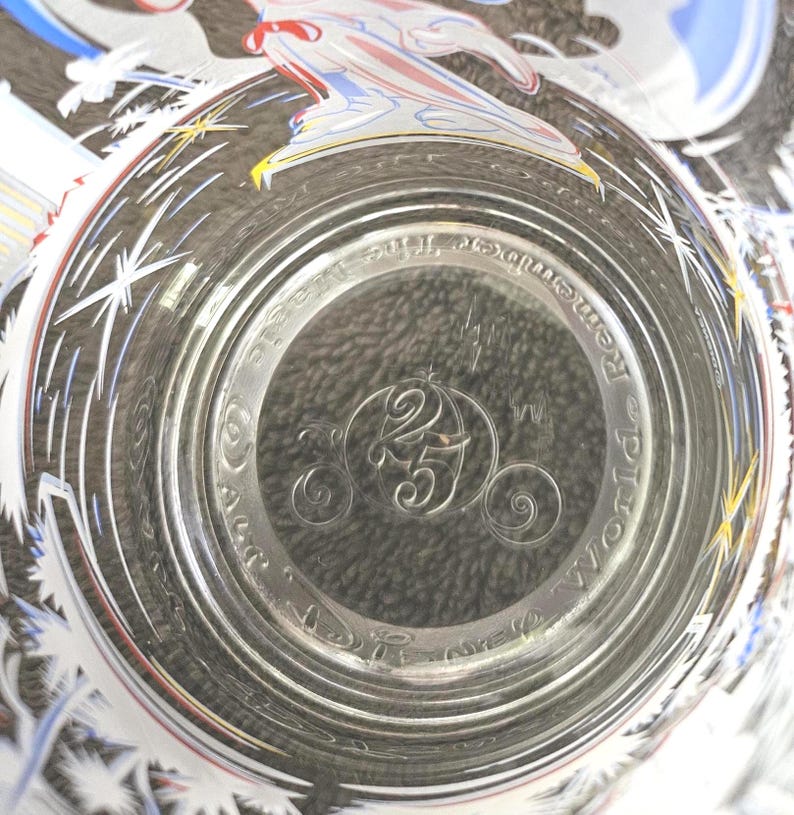 May include: Clear glass with a decorative rim featuring white starbursts and a colorful design. The bottom of the glass has the text "Remember the Magic" and a carriage with the number "25" inside.