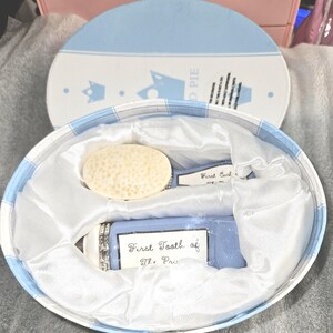 May include: A baby keepsake set in a blue and white oval box. The set includes a blue container labelled "First Tooth of The Prince", a small comb, and a round, textured object. The box has a blue and white gingham pattern.