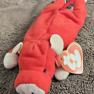 May include: Red plush toy bull with a white face and white ears. The toy has a red tail and a Ty tag attached to its ear.