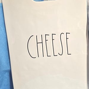 Puede incluir: Tabla de queso rectangular blanca con asa, con la palabra "CHEESE" en letras negras, estilo manuscrito. La tabla tiene una superficie lisa y un asa recortada en la parte superior. El fondo es una mezcla de azul y blanco.