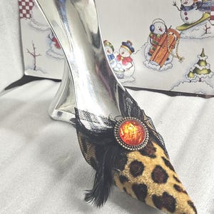 May include: A decorative silver high heel shoe with a leopard print toe, black lace, and a red and orange jewel. The shoe has a silver heel and is set against a winter-themed background.