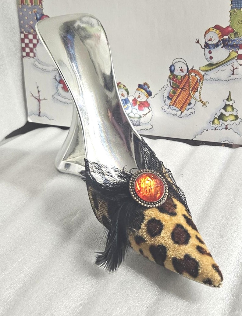 May include: A decorative silver high heel shoe with a leopard print toe, black lace, and a red and orange jewel. The shoe has a silver heel and is set against a winter-themed background.