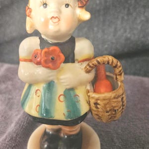 Op de afbeelding: Vintage keramieken beeldje van een jong meisje met blond haar, gekleed in een jurk en met een mandje. Het beeldje heeft een gedetailleerd gezicht en is geschilderd met zachte kleuren. Het mandje bevat een kleine fles.