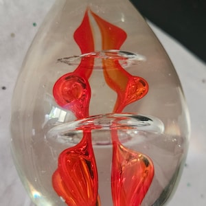 Può includere: Un fermacarte in vetro trasparente con un vortice rosso e arancione all'interno. Il vortice ha la forma di una fiamma e ha alcune bolle.