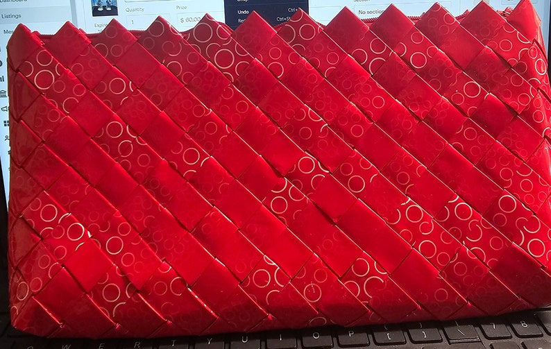 K&ouml;nnte beinhalten: Eine rote Clutch mit gewebtem Design. Die Clutch besteht aus rotem Material mit einem sich wiederholenden Muster aus wei&szlig;en Kreisen. Das gewebte Muster erzeugt eine strukturierte Oberfl&auml;che. Die Clutch ist rechteckig und hat einen Rei&szlig;verschluss.