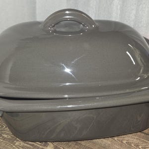 May include: A gray ceramic casserole dish with a lid. The dish has a rounded shape with a handle on the lid. The dish is sitting on a wooden surface.