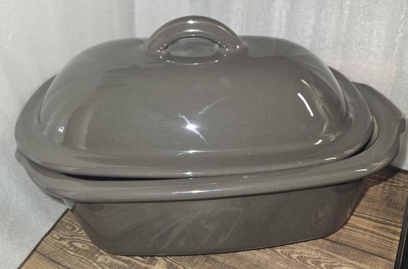 May include: A gray ceramic casserole dish with a lid. The dish has a rounded shape with a handle on the lid. The dish is sitting on a wooden surface.