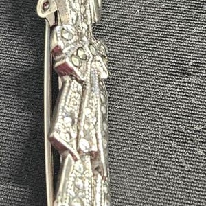 Pode incluir: Broche prateado retratando uma figura em um vestido longo, segurando um pequeno objeto. O broche tem detalhes intrincados e um fecho de alfinete na parte de trás. O vestido da figura tem um padrão texturizado, em um fundo escuro.