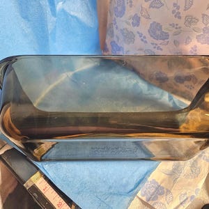Könnte beinhalten: Eine rechteckige, klare Backform aus Glas mit dunkler Tönung. Die Form hat eine leicht gebogene Form und Griffe an jeder Seite. Ein Maßband ist im Vordergrund zu sehen, das eine Größe von etwa 5 cm anzeigt.