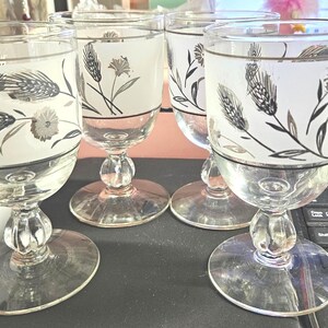 Op de afbeelding: Set van vier vintage glazen kelken. Elke kelk heeft een mat wit ontwerp met zilveren en zwarte tarwe- en bloemmotieven. De kelken hebben een heldere, ronde basis en een decoratieve steel.