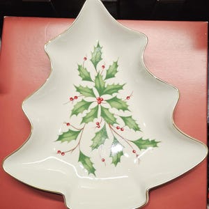 Può includere: Un piatto da portata in ceramica bianca a forma di albero di Natale con un bordo dorato. Il piatto presenta un ramo di agrifoglio verde con bacche rosse.