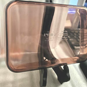 Puede incluir: Una fuente rectangular Pyrex, transparente y teñida de rosa. El plato tiene esquinas redondeadas y un borde. El texto "PYREX" y otros detalles son visibles. Está sostenido por un soporte negro. Capacidad de 750 ml.