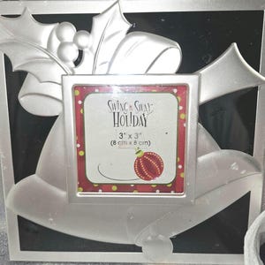 Puede incluir: Marco de fotos plateado en forma de campana con un diseño de rama de acebo. El marco tiene una abertura de 7,6 x 7,6 cm y dice "Swing & Sway Holiday" en la parte delantera.