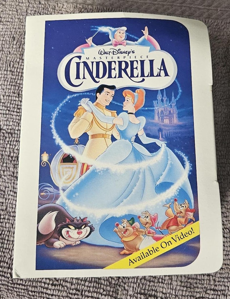 May include: A vintage VHS tape case featuring a scene from the Disney movie Cinderella. The case is white with a blue background and features Cinderella and Prince Charming dancing. The text "Walt Disney's Masterpiece Cinderella" is printed in blue and white. The text "Available On Video!" is printed in yellow on a red banner.