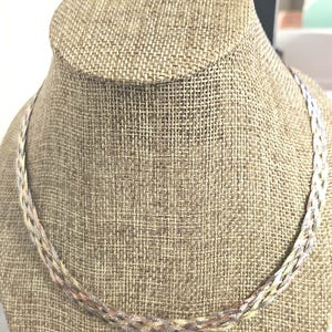 May include: A braided necklace with a mix of silver, gold, and rose gold tones. The necklace is displayed on a tan mannequin head. The chain is made of interwoven metal strands, creating a textured and elegant look. The necklace is a delicate piece of jewelry.