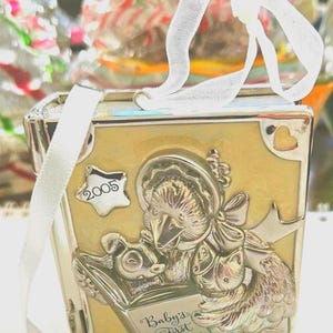 Può includere: Ornamento natalizio argentato di forma rettangolare con sfondo giallo chiaro. L'ornamento presenta un rilievo dettagliato di un uccello che legge un libro con la scritta "Baby's First Christmas". Un nastro bianco è attaccato per appenderlo e l'anno "2005" è visualizzato.