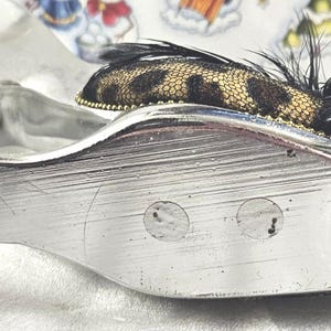 May include: A silver-toned, shoe-shaped trinket dish with a decorative leopard print and feather accent. The dish has a curved design and a textured surface, with a small leopard print fabric and black feathers.