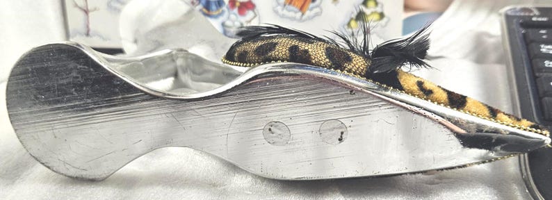 May include: A silver-toned, shoe-shaped trinket dish with a decorative leopard print and feather accent. The dish has a curved design and a textured surface, with a small leopard print fabric and black feathers.