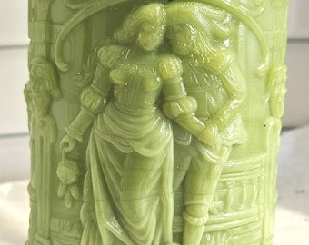 Puede incluir: Una jarra de vidrio verde con un relieve detallado de una pareja con ropa de época. La jarra tiene un borde y una base decorativos, y el relieve representa a un hombre y una mujer en un abrazo romántico. El diseño general es vintage.