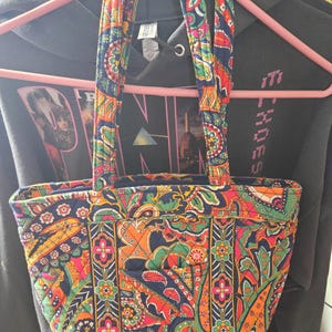 Può includere: Una borsa tote colorata con un motivo paisley nei toni del blu, arancione, verde e rosa. La borsa ha due manici e una forma rettangolare. La borsa è appesa a una gruccia rosa.