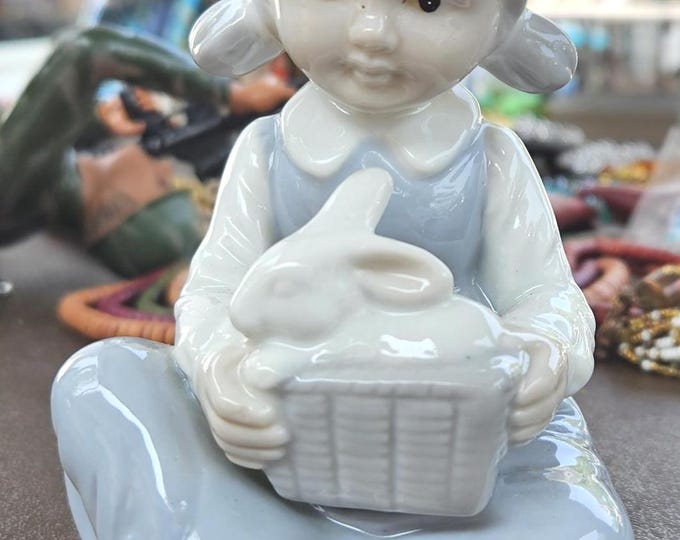 A vintage young lady is sitting on the ground holding her pet bunny in a basket. C7/7