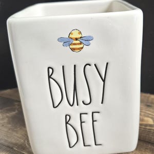 Puede incluir: Contenedor cuadrado de cerámica blanca con una ilustración de abeja de dibujos animados y las palabras "BUSY BEE" en letras negras. La abeja es amarilla y marrón con alas azules. El contenedor está sobre una superficie de madera.