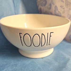 Puede incluir: Un cuenco de cerámica blanco con la palabra "FOODIE" en letras negras, de estilo manuscrito. El cuenco tiene forma redondeada y se asienta sobre una pequeña base circular. El fondo es una superficie azul claro.