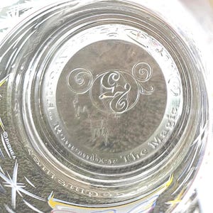 May include: Clear glass with etched design. The bottom of the glass features the Disney logo with the number 25 and the words "Remember The Magic". The glass has a starburst pattern on the side.