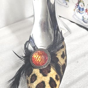May include: A decorative shoe-shaped bottle stopper. The shoe features a leopard print design on the toe, black lace detailing, and a red jewel. Black feathers add a touch of elegance. The stopper is silver.