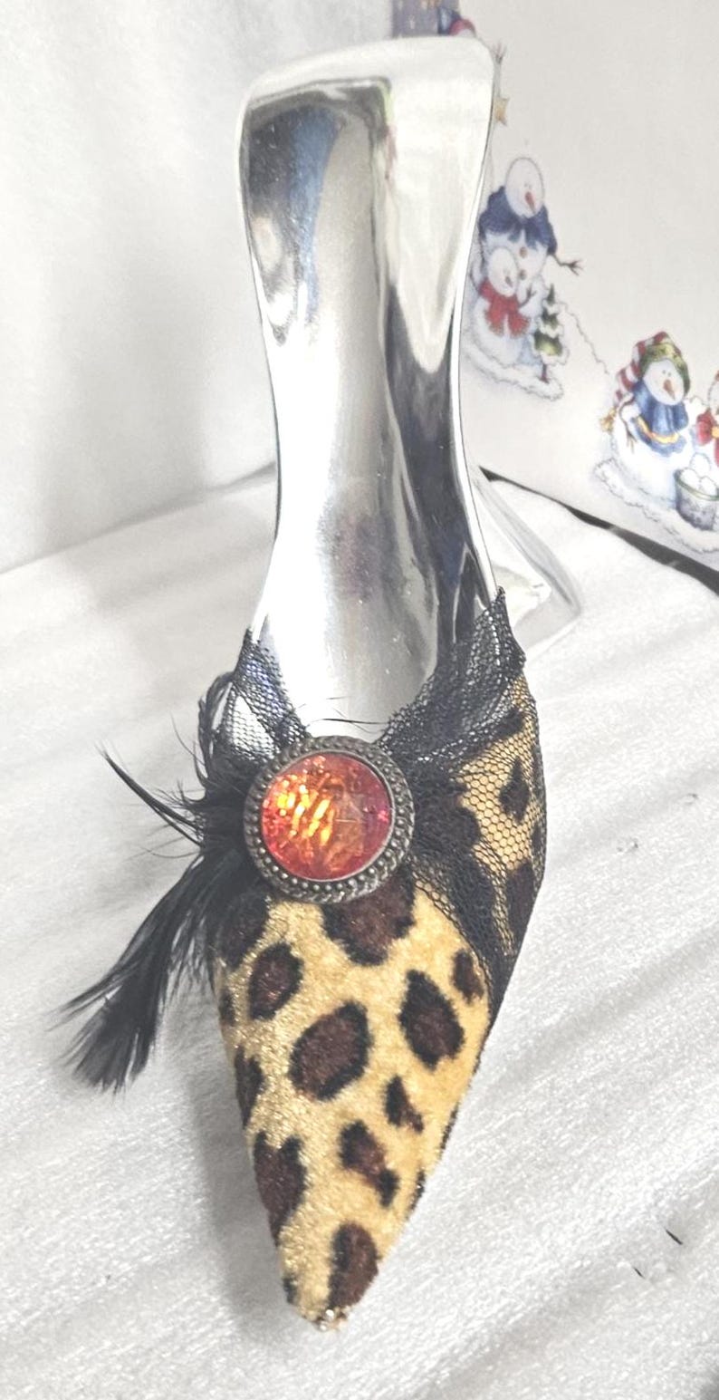May include: A decorative shoe-shaped bottle stopper. The shoe features a leopard print design on the toe, black lace detailing, and a red jewel. Black feathers add a touch of elegance. The stopper is silver.