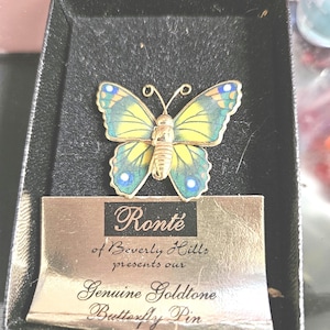 Puede incluir: Un broche de mariposa dorado con alas amarillas y azules, presentado en una caja negra. El broche está etiquetado como "Ronté of Beverly Hills" y se describe como un "Genuine Goldtone Butterfly Pin."